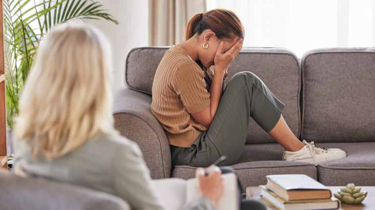 A young woman sits on a sofa with her head in her hands while another woman sits writing in a notebook.