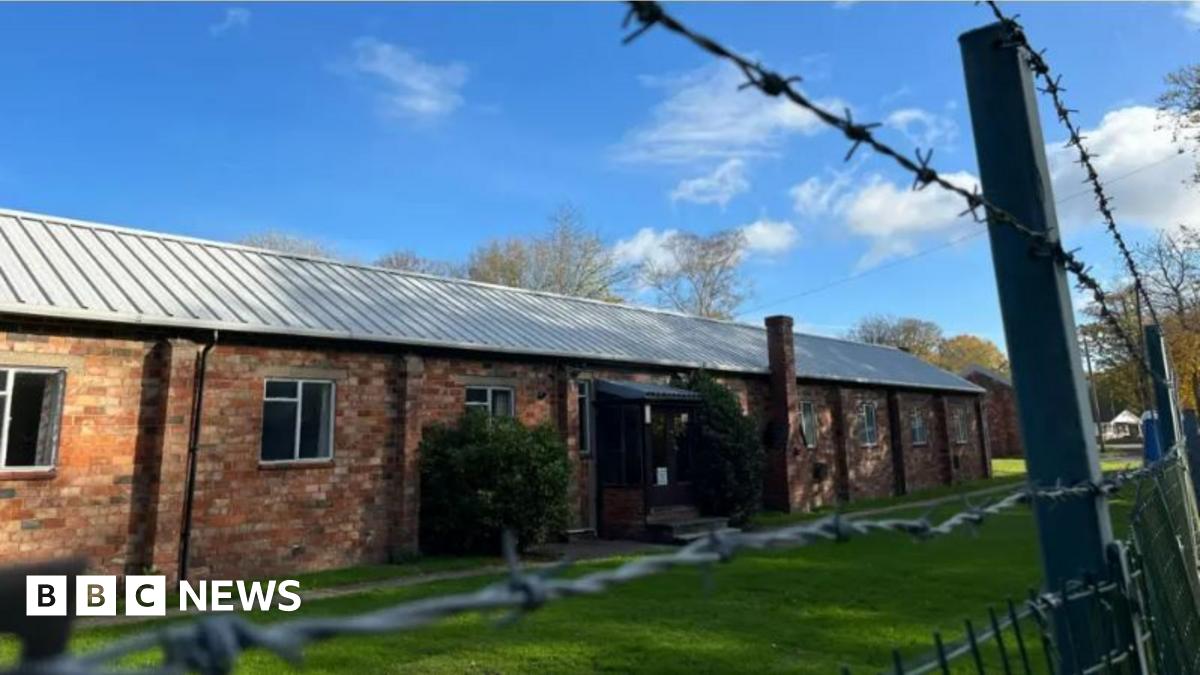 A brick building. It is sunny. Barbed wire is in front of it.