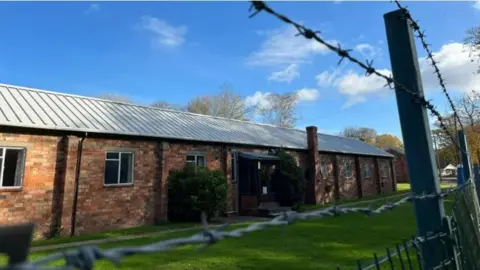 BBC A brick building. It is sunny. Barbed wire is in front of it.