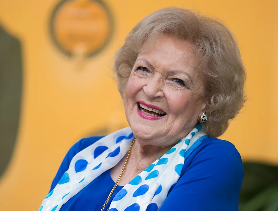 Betty White with curly hair wears a polka dot scarf and blue attire, exuding warmth and charisma