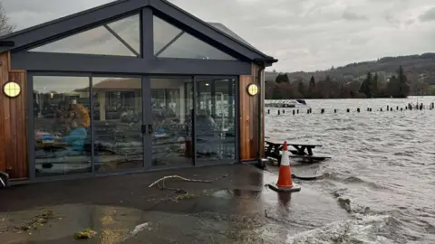 AMY DIXON Coniston Water lapping against the building of Bluebird Cafe in Coniston. The flooding is flush against the building. The lake is flooded and there are heavy grey rain clouds in the sky. People can be seen seated inside through the large glass doors at the gable end of the building.