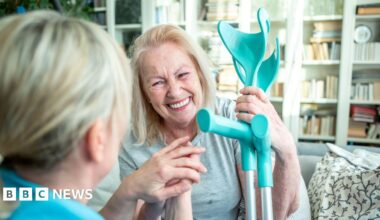 A healthcare worker with long blonde hair holding the hand of an older lady with crutches in her hair. She also has blonde hair and is wearing a grey t-shirt.
