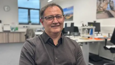Huw Watkins An office environment with a man seated in the foreground wearing a dark collared shirt. Behind him and slightly blurred, two people are working at desks with computer monitors. The room has white walls, a large window letting in natural light, and office furniture including chairs and storage units. A framed picture is partially visible on the wall to the left.