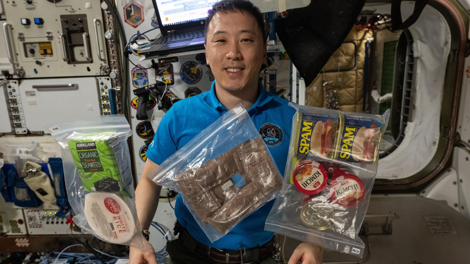 A man with short black hair wearing a dark blue shirt floats in microgravity behind various plastic covered food packages.