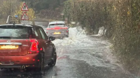 The Lake District Foundation A flooded road at Newby Bridge, Cumbria. There two cars - one red and one grey - that are travelling down the road where there is a significant amount of water covering the road. 