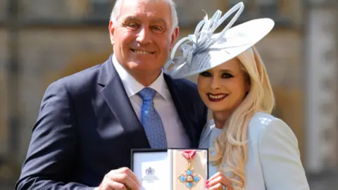 Getty Images Peter Shilton is dressed in a navy blue suit, a blue tie and a white shirt. Steph Shilton is wearing a light blue hat, and a light blue blazer. They are looking at the camera smiling, while holding a medal in a case.