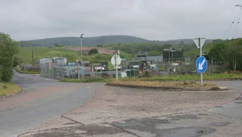 Richard Sutcliffe A council recycling site viewed from a distance with a fence round it and many containers inside