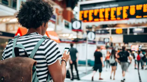 Getty Images Commuter stands with phone in hand at a busy railway station.
