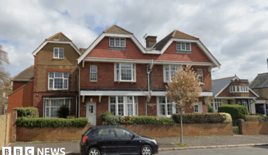 A large three-storey building made of red and yellow bricks, with a roof pointed in three places and large, white windows. A small black car is parked on the road in front of the property.