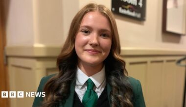 A young woman with long brown hair and a green school uniform on with green tie and a white blouse and green blazer
