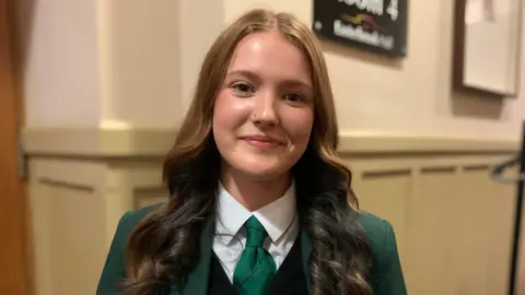 BBC A young woman with long brown hair and a green school uniform on with green tie and a white blouse and green blazer