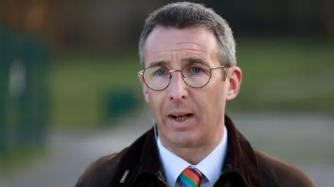 PA Media Muir is speaking. He wears glasses. He has a brown coat on with a white shirt and a colourful tie. 