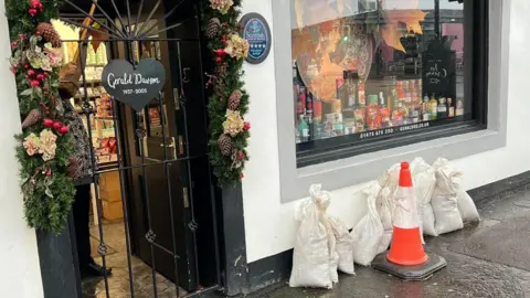 Geraldo's The front of a shop. Sandbags and a traffic cone are placed against the front wall to try and protect it from the rain.