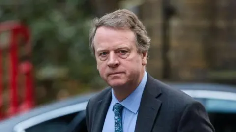 PA Media Alister Jack - a man with greying fair hair, wearing a dark suit with a blue shirt and patterned tie - pictured shortly after getting out of a car in London