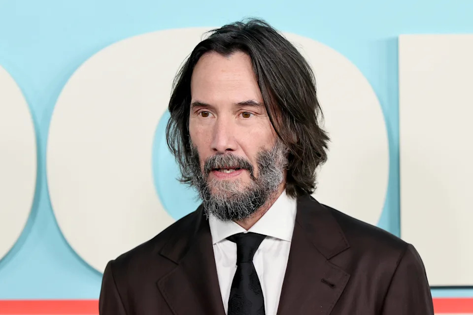 Keanu Reeves with long hair and a beard wearing a brown suit and black tie at a formal event