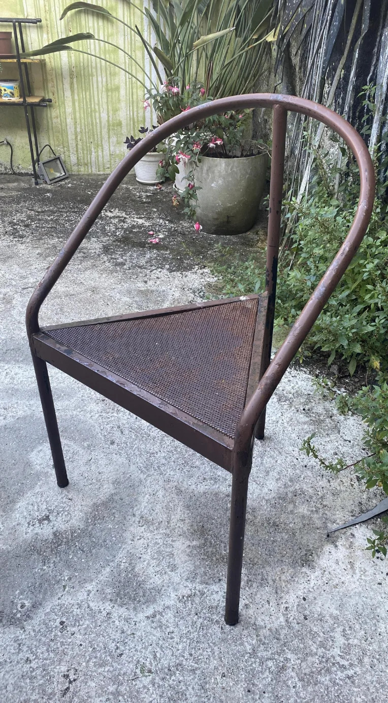 Metal chair with a minimalist, triangular seat design, placed on an outdoor patio surrounded by plants