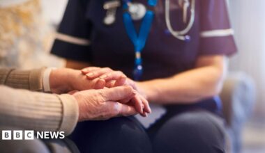 A nurse holds the hand of an elderly patient. The nurse is wearing blue scrubs and wears a blue lanyard and a pocket watch on her uniform. The elderly patient has a band around their wrist and wears a beige jumper. Neither of their faces are visible.