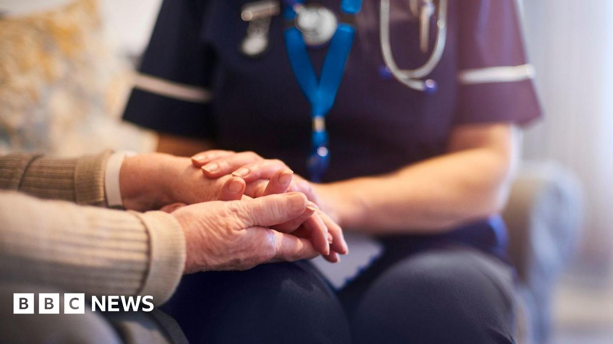 A nurse holds the hand of an elderly patient. The nurse is wearing blue scrubs and wears a blue lanyard and a pocket watch on her uniform. The elderly patient has a band around their wrist and wears a beige jumper. Neither of their faces are visible.
