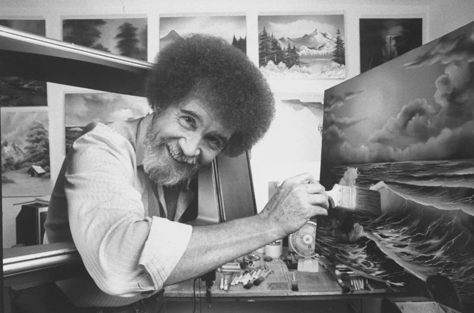 Bob Ross smiling while painting a seascape in a studio filled with landscape artworks
