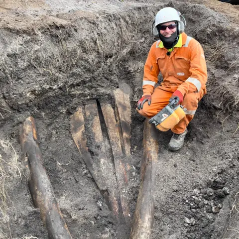 Paul White kneels beside part of the skull which is partly uncovered in the ground, the rest is covered by soil. He is wearing an orange boiler suit and a hard hat smiling at the camera.