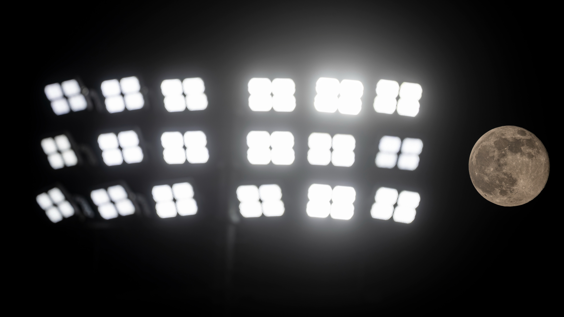A grey full moon is pictured shining to the right of a bank of stadium floodlights against a black night sky.
