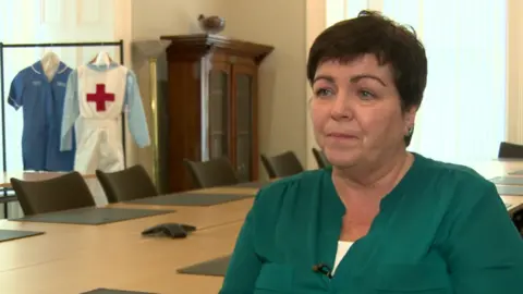 A head shot of Rita Devlin. She has short brown hair. She is wearing a green blouse with a white top underneath.