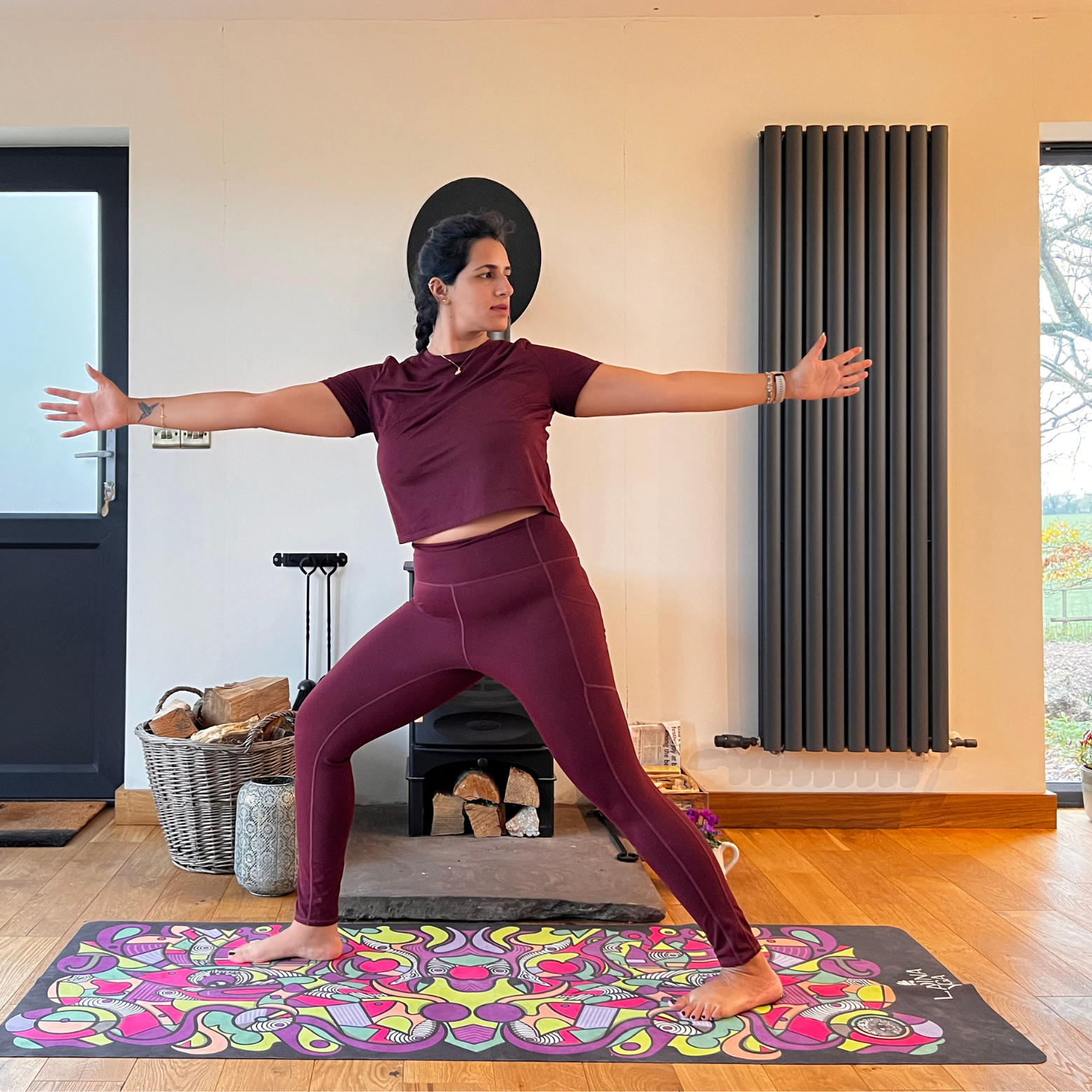 Maysun Hassanaly stands on a yoga mat performing revolved warrior. She is in a lunge position, with her right foot forward, right knee bent and left leg extended behind. Her torso is twisted to face left and her arms are extended at shoulder height.