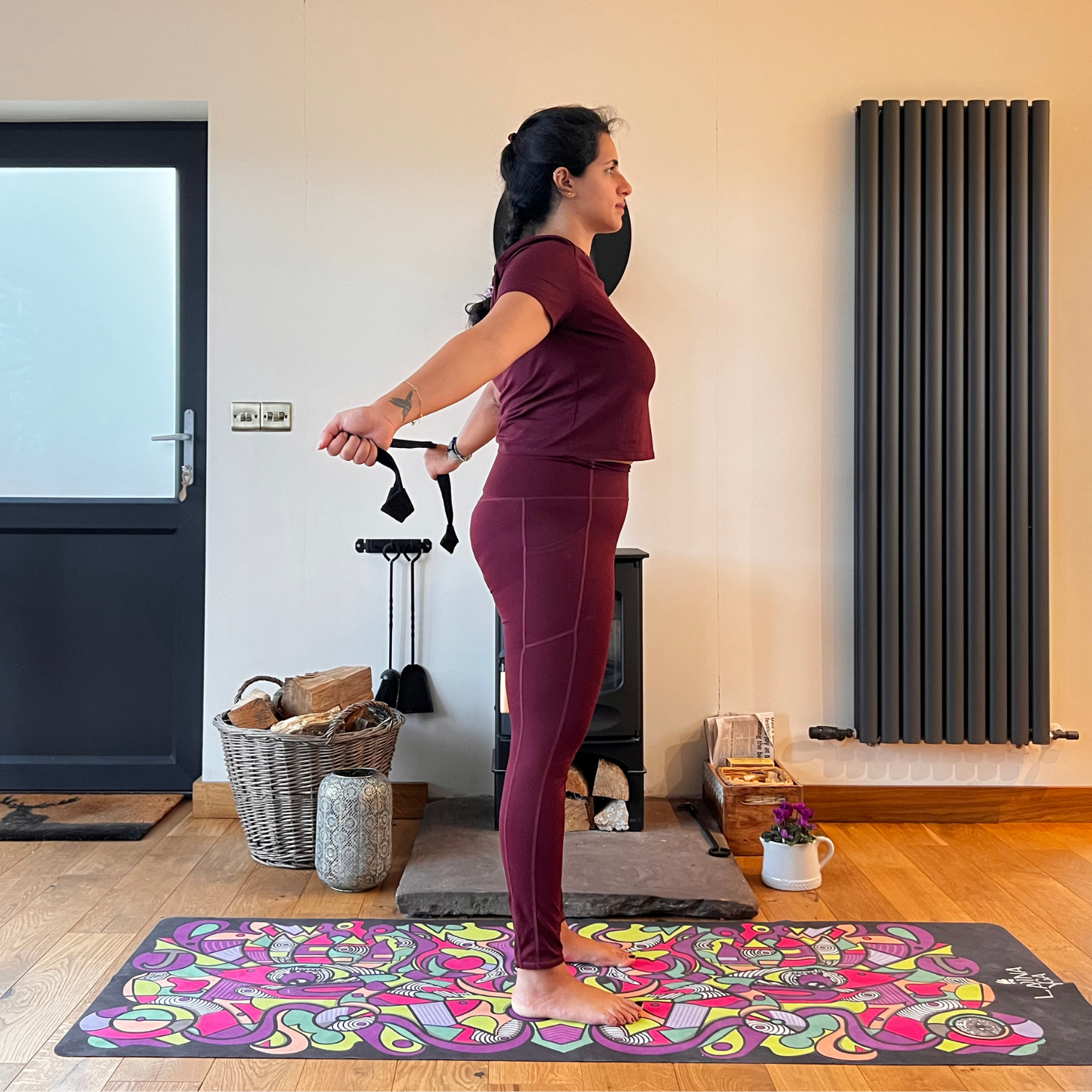 Maysun Hassanaly performs a shoulder floss movement with a yoga strap. She is standing on a colorful yoga mat, holding the strap behind her lower back with arms wide.