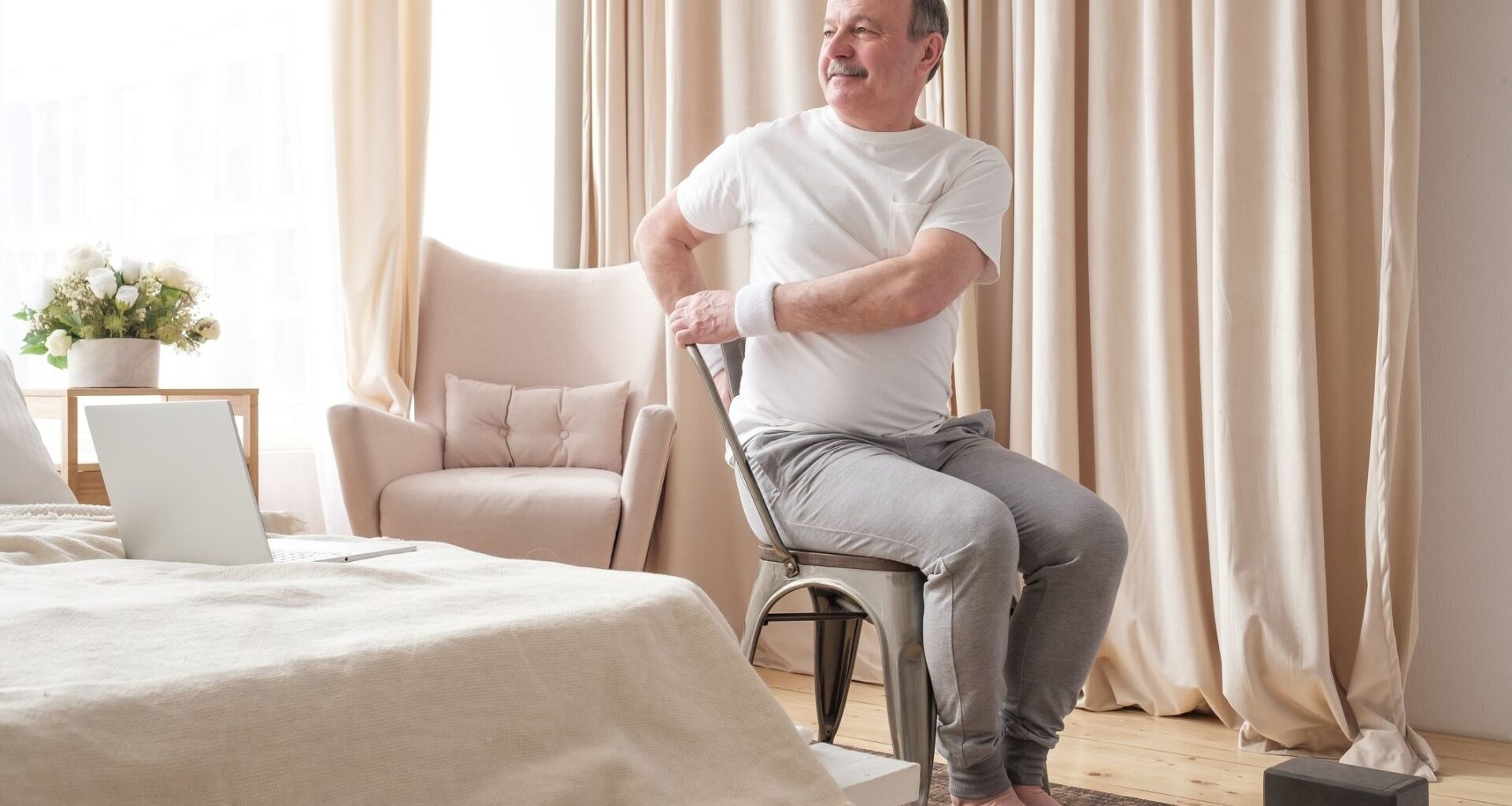 man in a white tshirt and grey joggers sitting in a chair and twisting to one side. he's in a bedroom setting with a laptop on the bed and a yoga block on the floor.