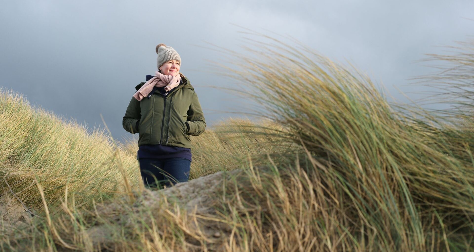 Woman wearing coat and hat walking through reeds on beach against stormy autumnal sky
