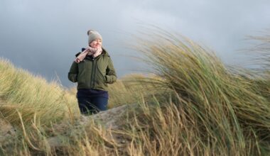 Woman wearing coat and hat walking through reeds on beach against stormy autumnal sky