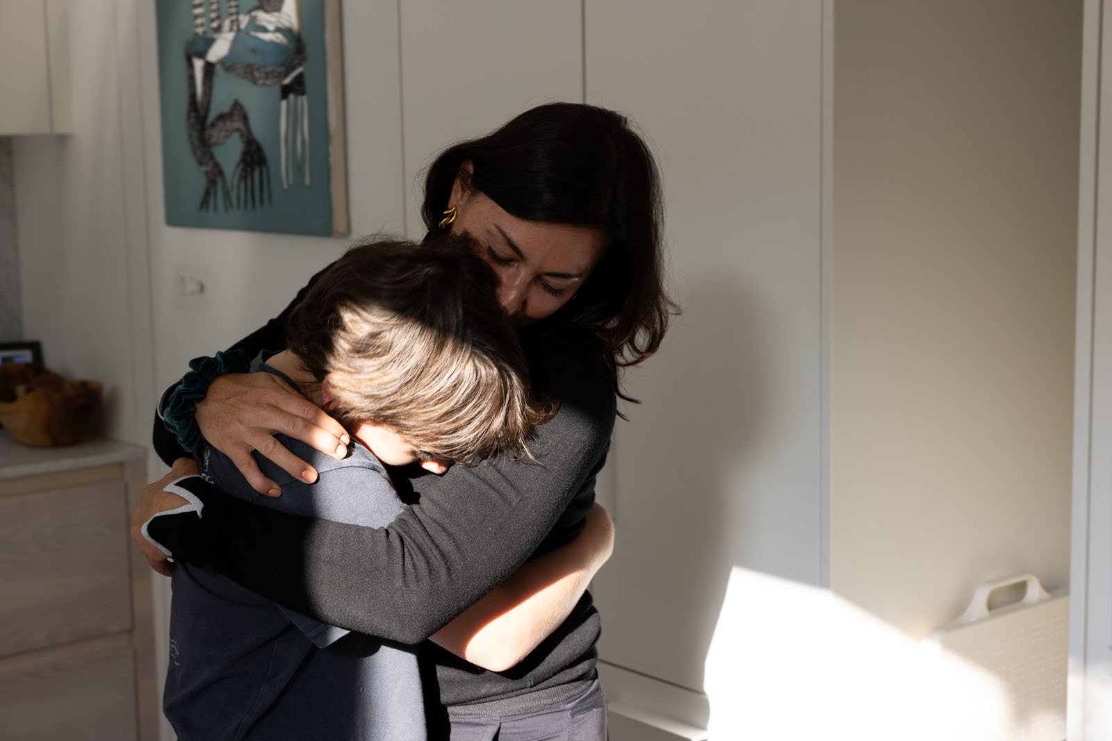 A woman and a child share a warm embrace in a sunlit room, both holding each other tightly. The woman’s eyes are closed, conveying a sense of comfort and support.