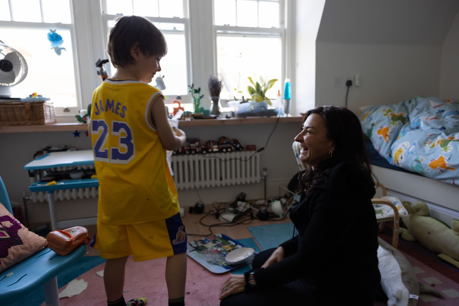 A young boy in a yellow LeBron James basketball jersey smiles at a woman sitting on the floor in a messy, sunlit bedroom decorated with toy cars, plants, and a bed with colorful sheets.