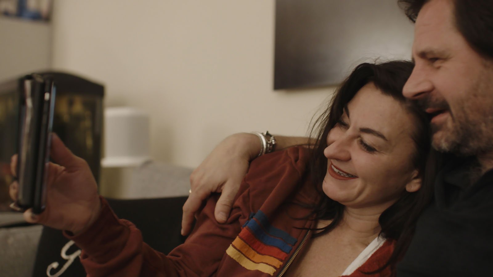 A woman and man sit closely together on a couch, smiling and taking a selfie or video call with a smartphone. The woman is wearing a red jacket with colorful stripes. The background is softly lit and out of focus.