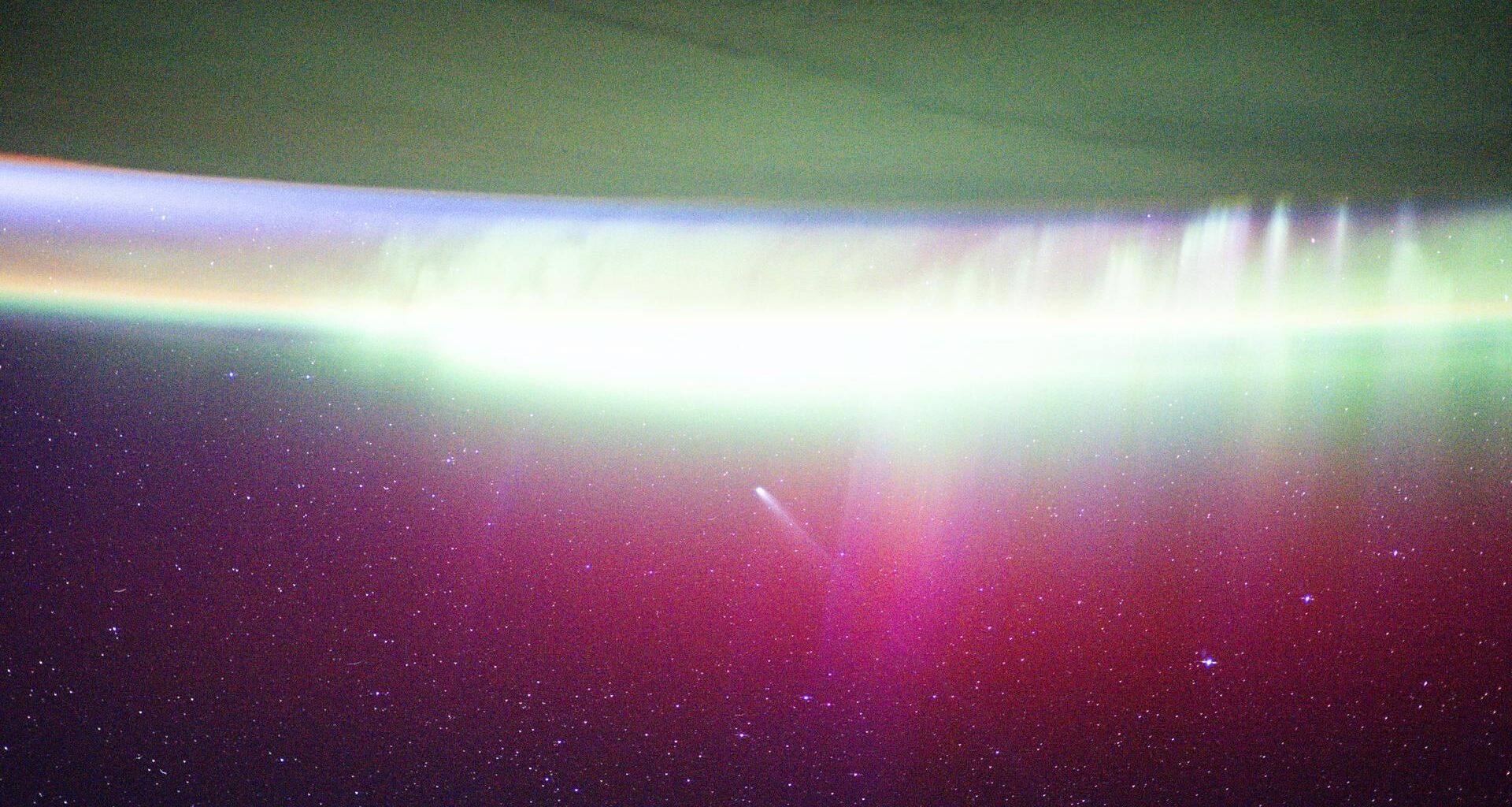 A streak of white showing Comet Lemmon moves into a red and green aurora seen on the edge of Earth in space