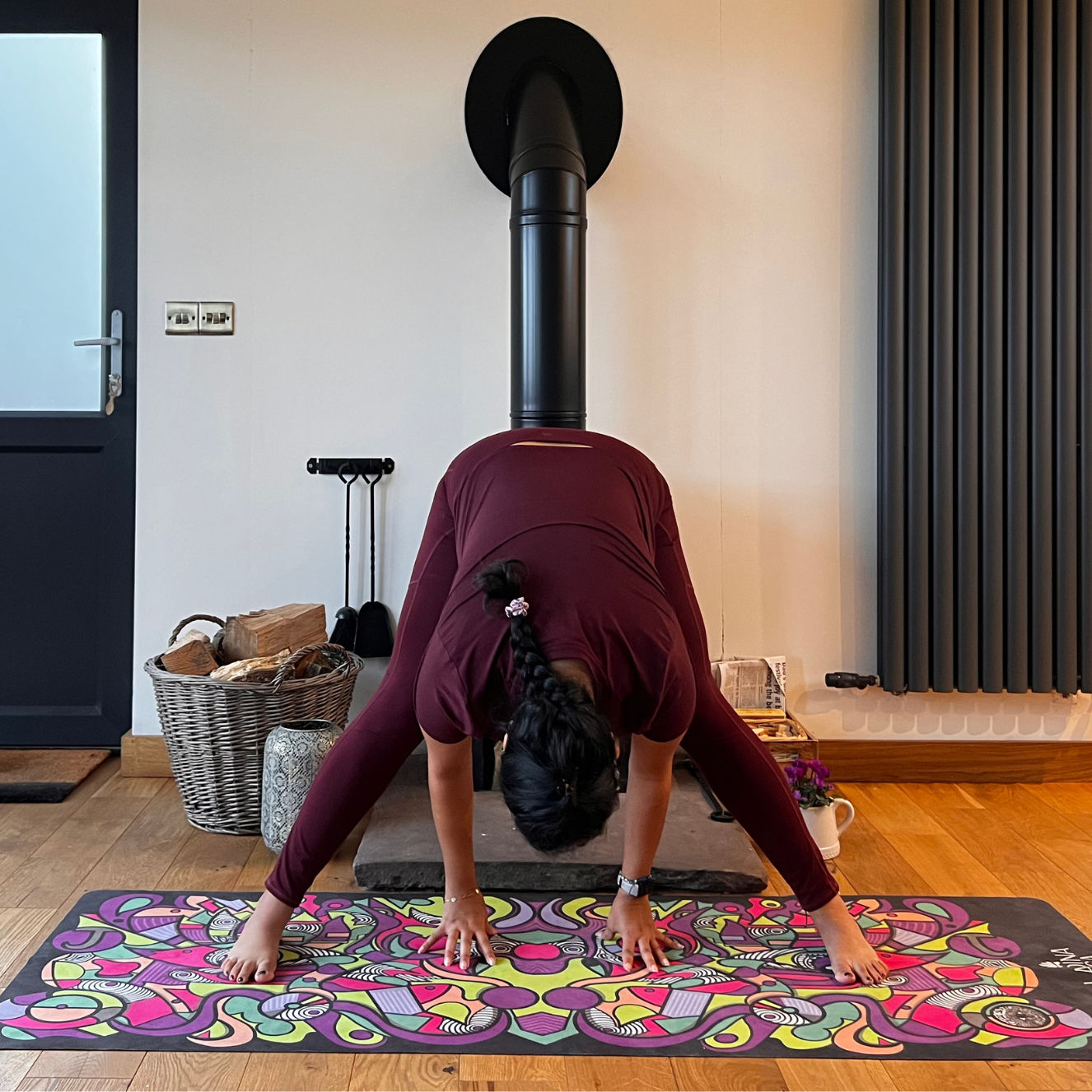Maysun Hassanaly performs a wide legged forward fold. She is standing on a colorful yoga mat with her legs wide apart, torso bent forward and hands on the floor.