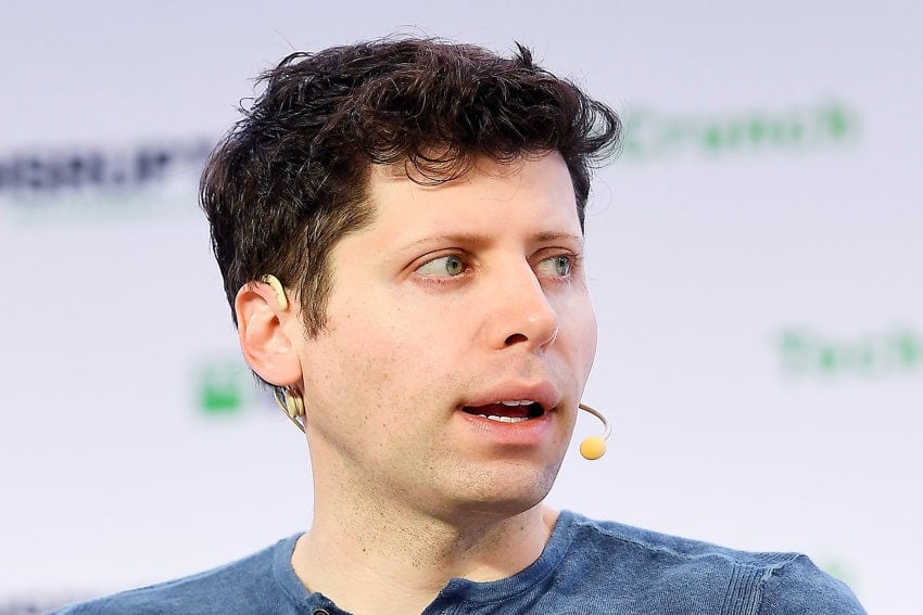 A close-up photograph of Sam Altman, head of the AI technology company OpenAI. He is wearing a small ear-mounted microphone and speaking at a public event.