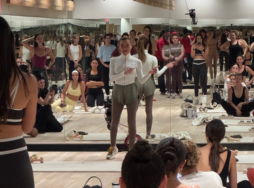 Photo of a fitness instructor leading a class in front of a studio mirror. Dozens of women in athleisure are seen in the mirror