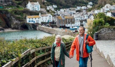 A senior man and his wife holding hands walking up a hill on a footpath looking away from the camera at the view. The fishing village of Polperro is behind them.