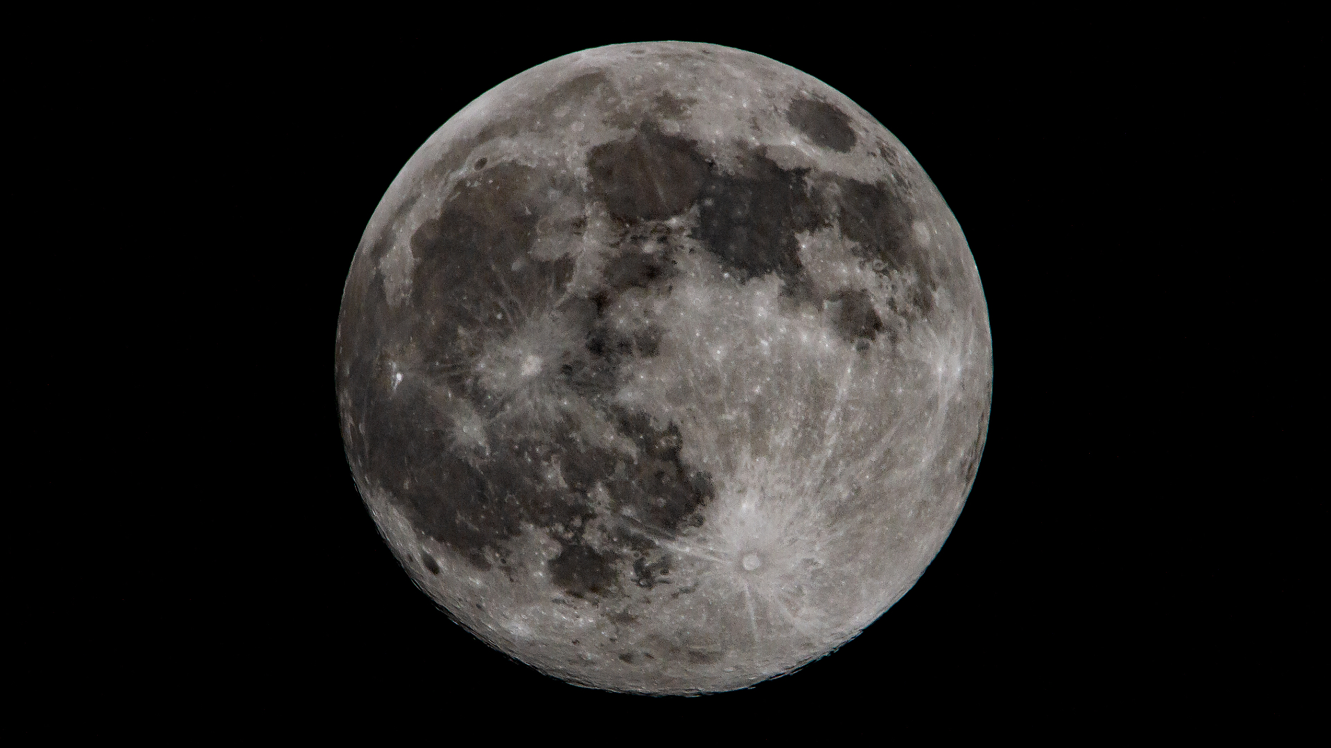 A full moon is pictured against a black night sky in sharp focus, revealing the presence of dark lunar seas and prominent craters from which bright material can be seen streaking across the lunar surface.