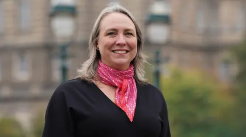Stewart Attwood Photography A woman with shoulder-length blonde-grey hair with a red and pink patterned scarf and a black V-neck jumper