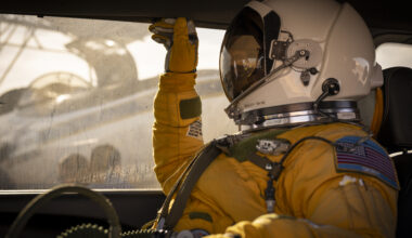 A person facing left sits inside a vehicle. They are wearing a yellow jumpsuit and gloves and a white helmet with a visor that looks like a smaller version of the kind astronauts wear during spacewalks. Outside of the window behind the person, you can see the ER-2 aircraft being prepared for flight.