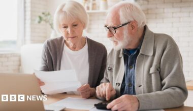Older man and woman sit at a table in a kitchen with paperwork and a laptop in front of them