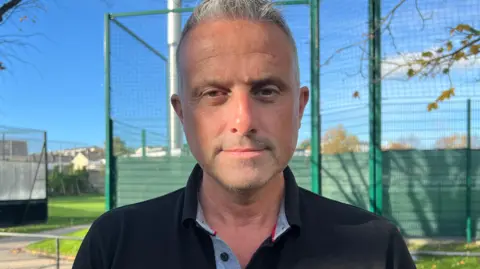 BBC Carl Walker is looking into the camera. He has short grey hair and is wearing a black polo shirt.