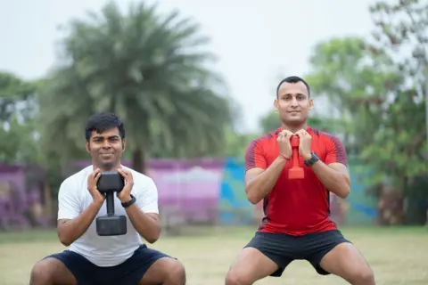 Getty Images Two men performing squats with dumbbells in an open outdoor environment. The image captures a moment of focused strength training, emphasizing dedication, fitness, and healthy lifestyle habits