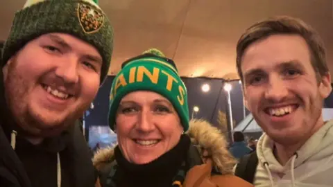 Family handout Three people - a smiling young man in green woollen hat on the left; a middle-aged woman in the centre in a green and yellow hat that says "Saints"; a young man on the right smiles with short brown hair.