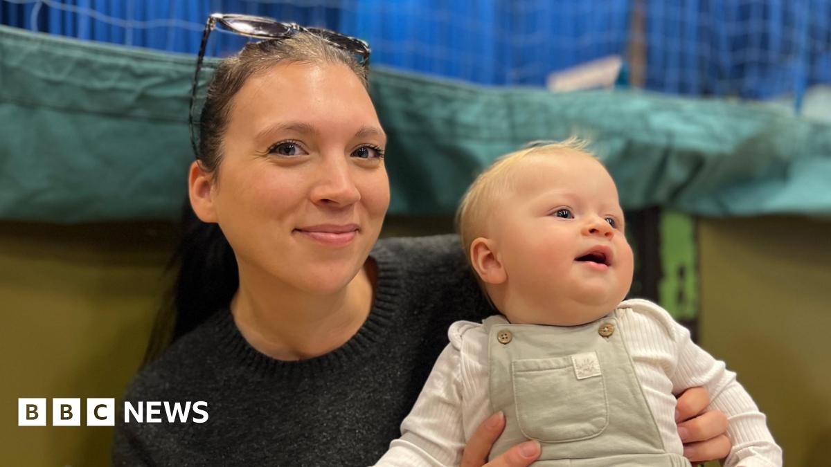 Leigh Walker and her baby Clyde - A woman with sunglasses on her head smiling at the camera. She's black hair in a ponytail and a black jumper. She is holding on her lap her baby who is wearing green dungarees and a white baby-gro. He's looking away from the camera and has a tuft of blonde hair on his wee head.