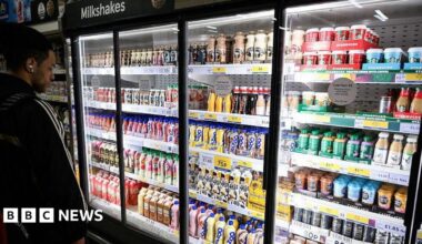A man with dark hair stands in front of a large supermarket fridge containing shelves of milk-based drinks, such as flavoured milkshakes and milky coffees like lattes, cappucinos and iced coffees.
