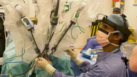 Mark Norman BBC A surgeon in an operating theatre  wearing scrubs and surgical head wear. They are monitoring three robotic arms that are operating on a patients throat cancer.  
