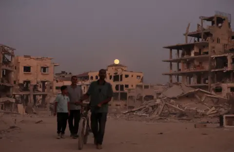 Dawoud Abu Alkas / Reuters Silhouetted figures walk in the foreground while the Moon rises over a building in the background, with a destroyed building and rubble to the right, in Gaza City on Wednesday.